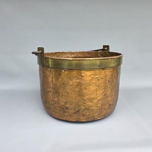 May include: A copper bucket with a hammered texture and a brass rim. The bucket has a rounded shape and two brass handles for carrying. The overall colour is a warm, aged copper tone, suggesting a vintage or antique item.