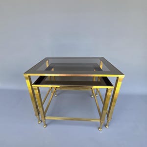 Peut inclure: Deux tables gigognes avec une structure en métal couleur laiton et des plateaux en verre foncé. Les tables sont de forme carrée, la plus petite s'insérant sous la plus grande. Les pieds sont fins et munis de boules.