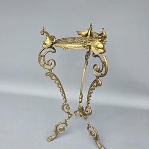 May include: A gold-colored, ornate plant stand with a round, patterned top. The stand features intricate scrollwork and decorative elements, including stylized floral or animal figures. The legs are curved and end in small, claw-like feet.
