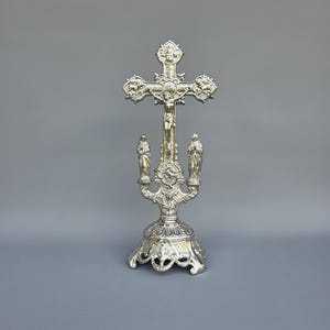 Pode incluir: Crucifixo prateado com um design detalhado. A cruz apresenta a figura de Jesus e é montada numa base decorativa com duas pequenas estátuas. A peça ornamentada é provavelmente utilizada para fins religiosos.