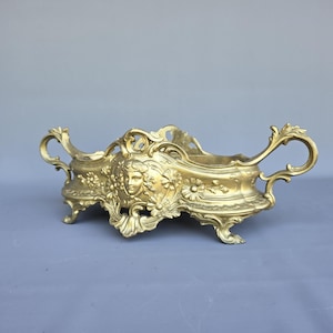 May include: A gold-toned metal centrepiece bowl featuring an ornate relief of a human face surrounded by floral motifs. The vessel has two curved handles on either side, a scalloped rim, and rests on four small, decorative feet with intricate scrollwork.