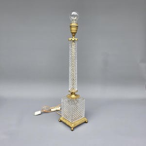 Puede incluir: Una lámpara de mesa de vidrio transparente con un patrón de diamantes en la base y la columna. La lámpara tiene una base y detalles metálicos dorados, y una bombilla en la parte superior. La lámpara está enchufada a una toma de corriente.