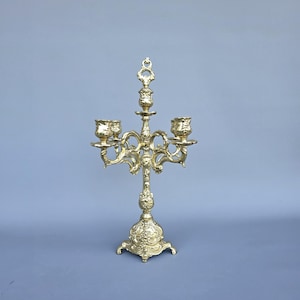 May include: A gold-colored candelabra with five candle holders. The ornate design features floral and scrollwork details. The base has a decorative, rounded shape with three feet. The candelabra is a decorative home accent.