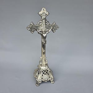 Op de afbeelding: Een zilverkleurig metalen crucifix dat op een sierlijke, opengewerkte voet met drie pootjes staat. Het kruis is voorzien van decoratieve krulranden en een centrale figuur van Jezus Christus met uitgestrekte armen. Het metaal heeft een gepolijste afwerking.