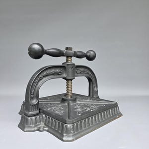 May include: A heavy, black cast iron book press featuring an arched frame with ornate floral relief detailing. The mechanism includes a central threaded metal screw and a horizontal handle with spherical ends for applying pressure to the flat base.