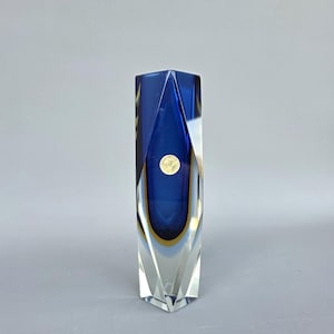 以下が含まれることがあります： 鮮やかな青と金の配色が印象的な、背の高いファセットガラスの花瓶。 透明なベースと濃い青色の中心部を持つ幾何学的なデザインです。 金色のバンドが青い部分を強調しています。 花瓶の高さは約25cmです。
