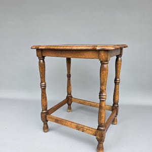 May include: A square wooden side table with a medium brown finish. The table features a flat top with slightly rounded edges and four turned legs connected by a rectangular lower support frame near the base, providing structural stability.