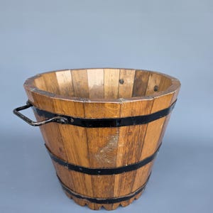 Op de afbeelding: Een houten emmer met een donkere metalen handgreep en zwarte metalen banden. De emmer is gemaakt van verticale houten latten en heeft een natuurlijke houten afwerking. De emmer is leeg en staat tegen een grijze achtergrond.