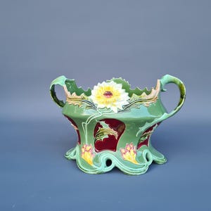 Op de afbeelding: Een groene keramische plantenbak met een bloemenontwerp. De plantenbak heeft twee handvatten en een geschulpte rand. De binnenkant is dieprood en de buitenkant heeft gele en witte bloemen.