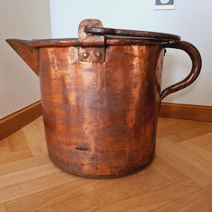 Op de afbeelding: Een grote, antieke koperen pot met een tuit en een gebogen handvat. De pot heeft een donkere, verouderde patina, die tekenen van gebruik vertoont. Het handvat is met klinknagels bevestigd. De pot staat op een houten vloer.