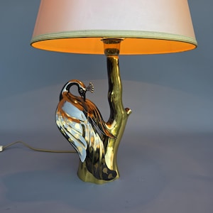 Op de afbeelding: Een messing pauwenlamp met een perzikkleurige lampenkap. De lamp heeft een gedetailleerd pauwenontwerp met zilveren en zwarte accenten, geplaatst op een messing tak. De lampenkap heeft een zachte perzikkleur.