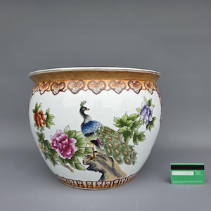 Op de afbeelding: Grote, ronde keramische bloempot met een witte basis, versierd met een kleurrijke pauw en bloemmotieven. De rand is warmbruin met decoratieve patronen. De pauw heeft blauwe, groene en bruine veren. De bloemen zijn roze, rood en paars.
