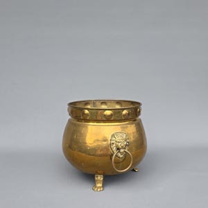 Pode incluir: Um vaso redondo de latão com uma faixa decorativa de tachas circulares em relevo na borda superior. O recipiente é sustentado por três pés em forma de garra e inclui uma alça de cabeça de leão na lateral com um anel circular preso.