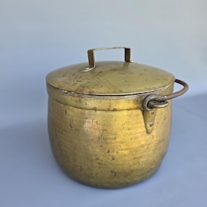 Może przedstawiać: Wintage miedziany garnek z pokrywką i uchwytem. Garnek ma zaokrąglony korpus i uchwyt z boku. Pokrywka ma uchwyt na górze. Mosiądz ma ciepłą, postarzaną patynę.