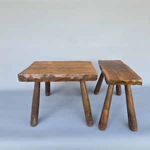 Puede incluir: Dos taburetes de madera rústicos. Uno es cuadrado, el otro rectangular. Ambos tienen tres o cuatro patas robustas e inclinadas. La madera tiene un tono marrón cálido natural, con un borde texturizado y tallado en la parte superior.