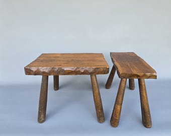 Puede incluir: Dos taburetes de madera rústicos. Uno es cuadrado, el otro rectangular. Ambos tienen tres o cuatro patas robustas e inclinadas. La madera tiene un tono marrón cálido natural, con un borde texturizado y tallado en la parte superior.