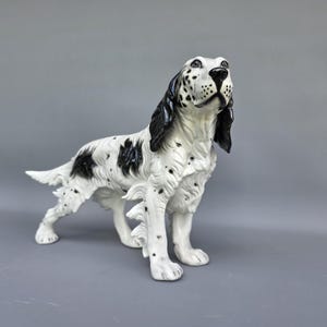 以下が含まれることがあります： 黒い斑点と模様がある白い毛並みの犬の陶器製フィギュア。犬は長く光沢のある黒い耳、黒い鼻、そして細かく質感のある毛並みが特徴です。頭を向けた状態で、警戒心のある立ち姿で表現されています。