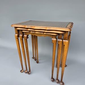 Puede incluir: Un juego de tres mesas nido de madera. Las mesas tienen un acabado marrón claro con un patrón tejido en la parte superior. Cada mesa tiene cuatro patas delgadas y curvas. La mesa más grande mide aproximadamente 61 cm de ancho.