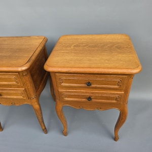 Op de afbeelding: Twee antieke houten nachtkastjes. Elk nachtkastje heeft twee lades met zwarte knoppen, gebogen poten en een lichtbruine afwerking. De bovenkanten zijn rechthoekig met afgeronde randen. De tafels zijn van hout.