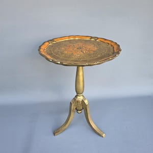 Può includere: Un tavolino rotondo con finitura dorata e bordo smerlato. Il piano del tavolo presenta un intricato motivo floreale in rilievo nei toni dell'arancione e dell'oro. Il tavolo poggia su una colonna centrale sostenuta da tre gambe curve.