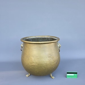 Könnte beinhalten: Ein großer, antiker Messingkessel mit rundem Körper und drei kleinen Füßen. Der Kessel hat einen breiten Rand und zwei Löwenkopfgriffe. Das Messing hat einen warmen, goldenen Farbton, und der Kessel scheint gealtert zu sein. Eine grüne und weiße Karte befindet sich neben dem Kessel.
