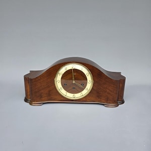 May include: A dark brown wooden mantel clock with a curved top and a circular clock face. The clock face features gold-coloured numerals and hands. The clock is made of wood and sits on a flat surface.