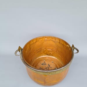 May include: An antique copper bucket with a rounded body and two metal handles. The interior has a golden hue, and the exterior shows signs of age. The rim is reinforced with a silver-coloured metal band.