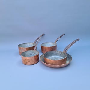 May include: Four copper saucepans with long handles are arranged on a light blue surface. The pans vary in size, with a small frying pan in the foreground. The copper has a warm, reddish-brown hue, and the interiors are a lighter, silvery color.