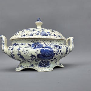 May include: A white and blue ceramic tureen with a lid. The tureen features a floral pattern in blue against a white background. It has two handles and sits on small feet. The lid is topped with a small finial.