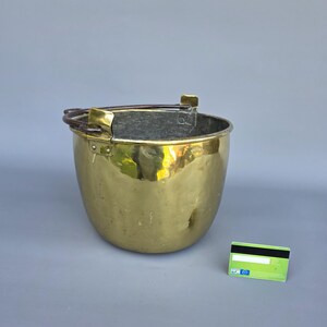 Op de afbeelding: Een glanzende messing emmer met een afgeronde vorm en een stevige handgreep. De emmer heeft een donkere binnenkant en is aan de naden geklonken. Een groene en witte creditcard ligt naast de emmer.