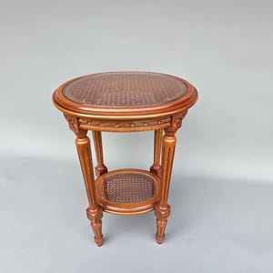 May include: A small, antique-style plant stand with a round top and a lower shelf. The stand is made of wood with a warm, reddish-brown finish. The top and shelf feature a woven cane design, and the legs are carved.