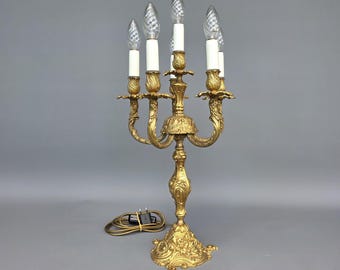 May include: A gold-toned metal candelabra lamp featuring five arms with white candle-style sleeves and clear, spiraled flame-shaped light bulbs. The base and central stem are decorated with ornate, raised floral and scrollwork patterns.