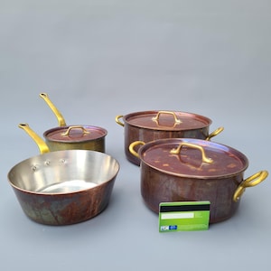 Op de afbeelding: Een verzameling koperen kookgerei met messing handgrepen. De set bevat een kleine steelpan, een koekenpan en twee potten met deksel. Het koper heeft een rijke, verouderde patina en de binnenkanten zijn zilverkleurig. Het kookgerei wordt op een grijze achtergrond gepresenteerd.