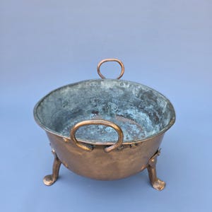 Op de afbeelding: Een koperen kom met een afgeronde vorm, voorzien van twee gebogen handvatten en drie kleine voetjes. De kom heeft een verweerde uitstraling met patina, met tinten bruin en blauwgroen. De binnenkant heeft een donkerdere, verouderde uitstraling.