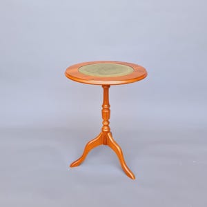 Op de afbeelding: Een kleine, ronde, houten bijzettafel met een driepoot. Het tafelblad heeft een cirkelvormige, olijfgroene inzet. De tafel is ongeveer 61 cm hoog.