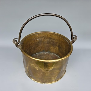 Op de afbeelding: Een vintage messing emmer met een gebogen metalen handvat. De emmer heeft een cilindrische vorm met een licht uitlopende rand. Het messing heeft een warme, verouderde patina. De handgreep is bevestigd met decoratieve lussen.