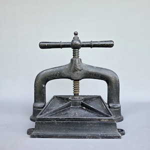 May include: A heavy, black cast iron book press featuring a central threaded screw mechanism with a horizontal handle bar. The base consists of a square, flat plate with reinforced structural ribs, designed for applying pressure to documents or books.