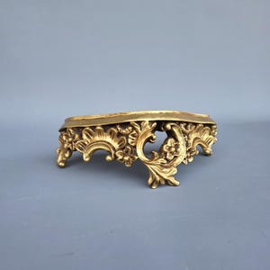 Op de afbeelding: Een goudkleurige, sierlijke decoratieve standaard met een gedetailleerd bloemen- en scrollontwerp. De standaard heeft een laag profiel met een platte bovenkant en decoratieve poten. De algehele esthetiek is vintage en elegant.