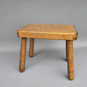 May include: A small wooden stool with a square seat and four rounded legs. The stool is made of light brown wood with visible grain, and the legs are slightly tapered. The design is simple and functional.