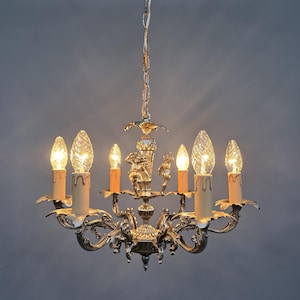 Può includere: Un lampadario d'argento con sei luci a forma di candela. L'apparecchio ha bracci ornati e curvi e figure decorative di cherubini. Le lampadine sono accese, proiettando un bagliore caldo. Il lampadario è appeso a una catena.