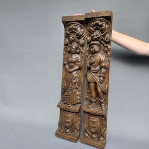 Peut inclure: Deux panneaux verticaux rectangulaires en bois brun avec des sculptures en relief détaillées. Chaque panneau représente un personnage en pied en tenue historique, surmonté d'un motif floral et d'un blason à la base.