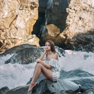 May include: A person wearing a white and black striped off-the-shoulder romper sits on a large rock near the ocean. The waves crash against the rocks, creating a dramatic scene. The rocks are a mix of gray and brown.