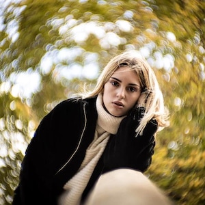 May include: A person with blonde hair wearing a black jacket and a cream-coloured turtleneck sweater. The background is blurred with bokeh effects of green and yellow foliage, creating a soft, artistic aesthetic.
