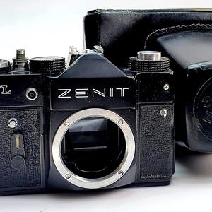 Puede incluir: Cámara Zenit TTL negra con montura de lente plateada y la palabra ZENIT impresa en la parte delantera. La cámara va acompañada de una funda de cuero negra. La cámara tiene varios diales y botones.