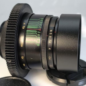 May include: A black camera lens with a gear-like focus ring and a lens hood. The lens has various markings and numbers, including aperture settings. The lens is lying on a white surface with a lens cap.