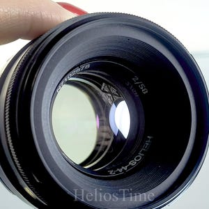 Puede incluir: Un objetivo de c&aacute;mara negro Helios-44-2 con el texto "Helios Time" en la parte inferior. El objetivo tiene una distancia focal de 58 mm y una apertura de f/2. El objetivo est&aacute; sostenido por una persona con esmalte de u&ntilde;as rojo.