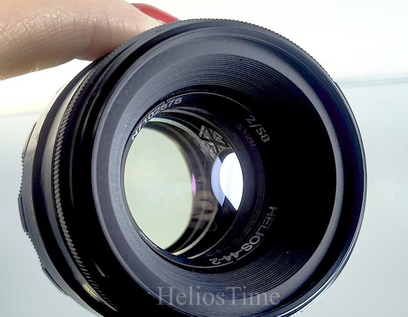 Puede incluir: Un objetivo de c&aacute;mara negro Helios-44-2 con el texto "Helios Time" en la parte inferior. El objetivo tiene una distancia focal de 58 mm y una apertura de f/2. El objetivo est&aacute; sostenido por una persona con esmalte de u&ntilde;as rojo.