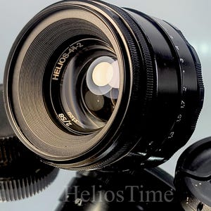 Puede incluir: Un objetivo de c&aacute;mara Helios-44-2 negro con un borde plateado y el texto "Helios-44-2 2/58". El objetivo va acompa&ntilde;ado de una tapa de objetivo y una tapa de cuerpo. Producto de fotograf&iacute;a.