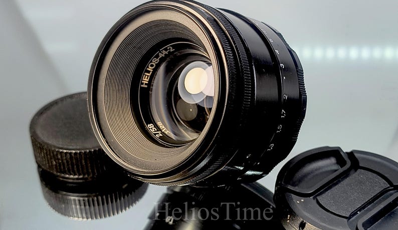 Puede incluir: Un objetivo de c&aacute;mara Helios-44-2 negro con un borde plateado y el texto "Helios-44-2 2/58". El objetivo va acompa&ntilde;ado de una tapa de objetivo y una tapa de cuerpo. Producto de fotograf&iacute;a.