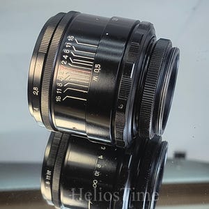 Puede incluir: Un objetivo de c&aacute;mara negro con marcas blancas y un anillo de enfoque estriado. El objetivo tiene una serie de n&uacute;meros y l&iacute;neas que indican los ajustes de apertura. El objetivo se refleja en una superficie espejada.